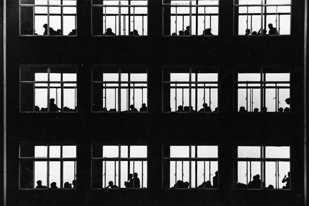 A monochrome photograph of a grid of classroom windows, students in silhouette