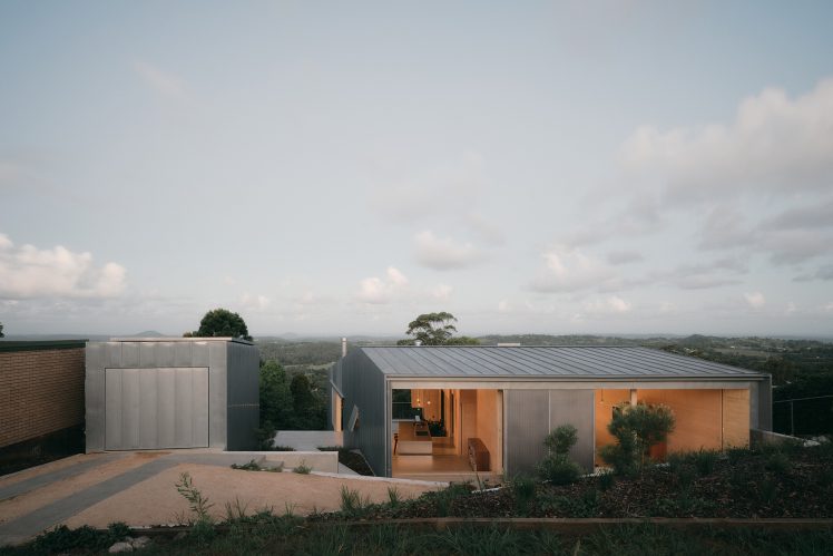 Atelier Chen Hung’s Mapleton House in Australia. Seen from the road at dusk with shutters open, letting out light from inside and showing the deep blue sky and the view to the valley below